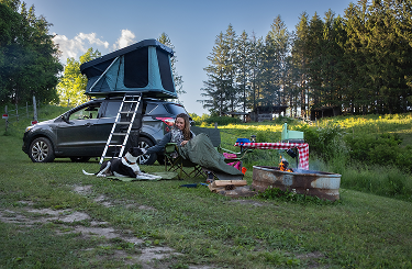 Woman  with a dog tucked beside her in a camp chair and an SUV with an open rooftop tent behind