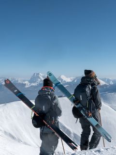 Two skiers with Thule backpacks and skis strapped to their backs walk through the snow.