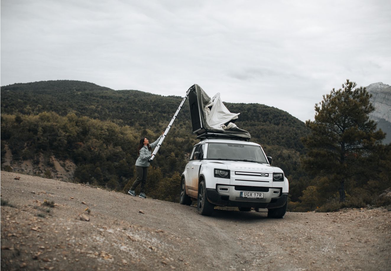 Человек раскладывает палатку Thule Approach, установленную на крыше автомобиля, припаркованного на горной дороге.