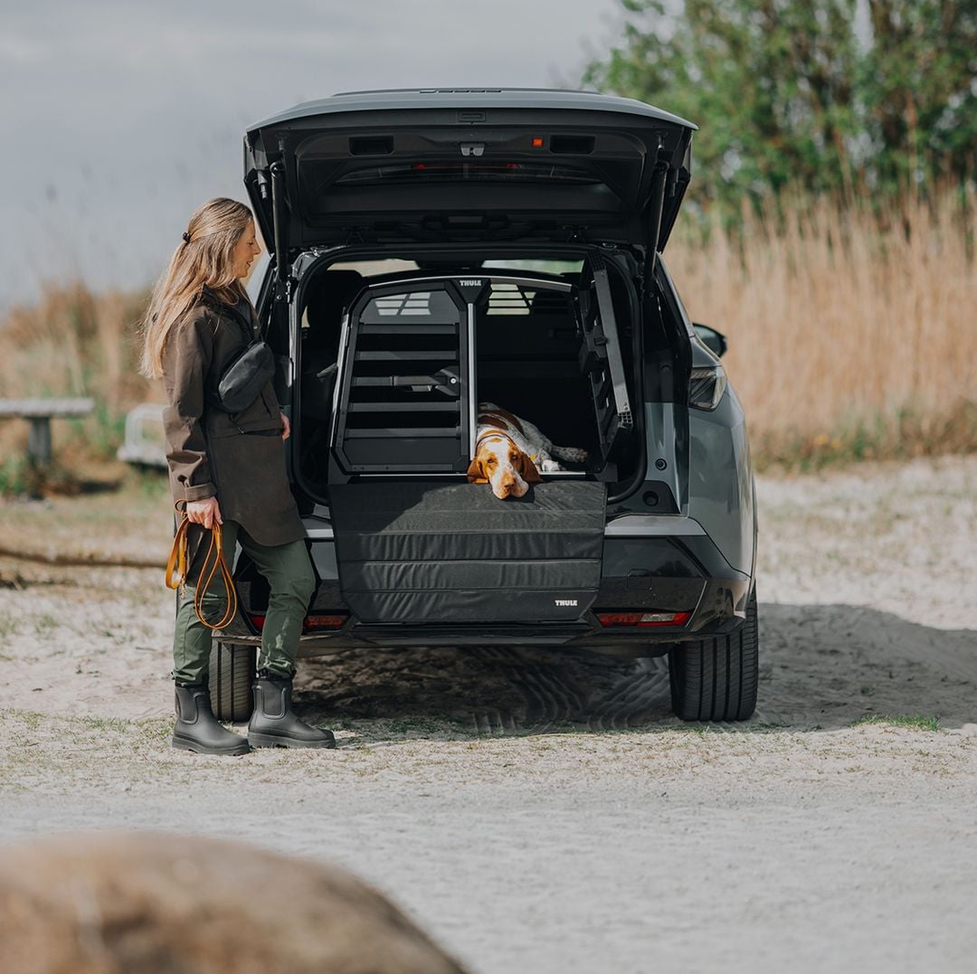 Kobieta stoi obok SUV-a z otwartym tylnym włazem, gdzie pies relaksuje się w klatce Thule Allax. Scena podkreśla bezpieczną i komfortową podróż psów w środowisku plenerowym.