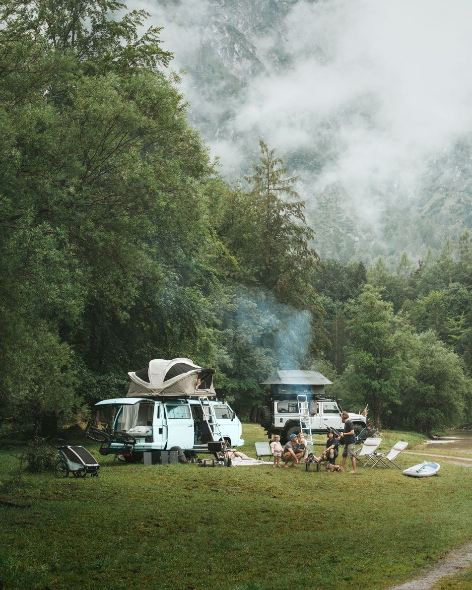 A van and Jeep with rooftop tents mounted to the top are parked at a campsite while people talk by a fire.