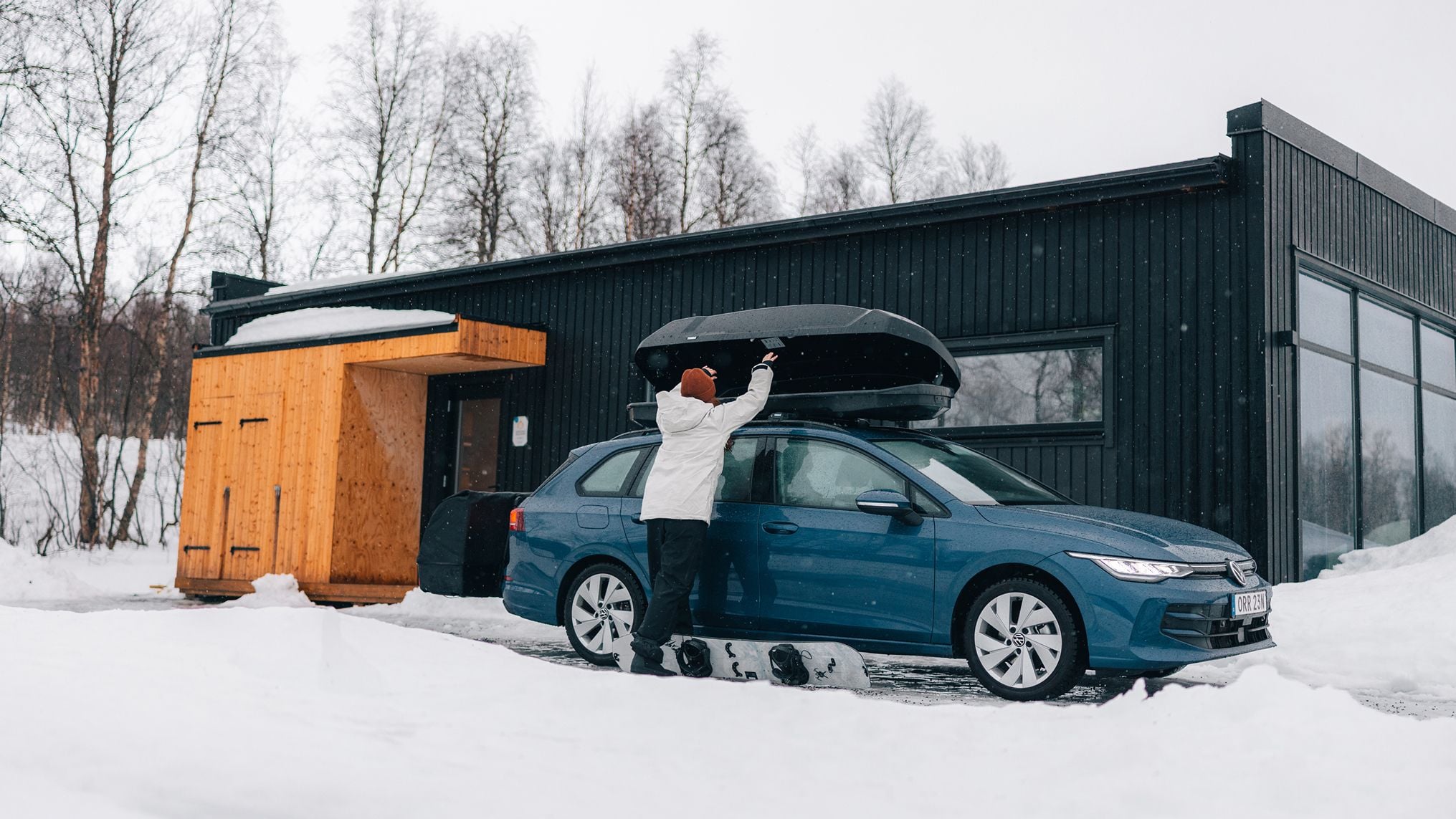 En person med en snowboard vid fötterna öppnar en Thule-takbox ovanpå en bil vid ett hus med snö runtomkring. 