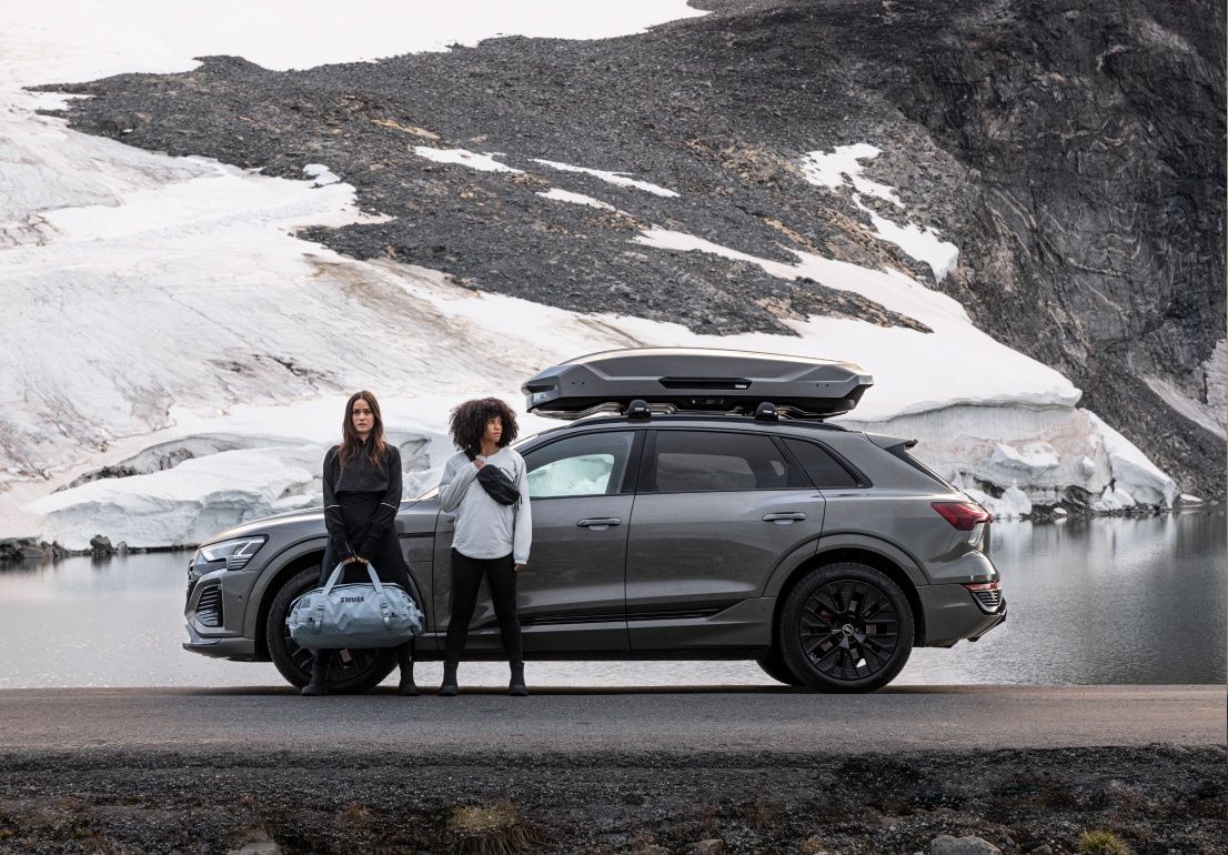 Two women, one holding a Thule duffel bag, stand in front of a gray SUV with a gray Thule Motion 3 roofbox on top.