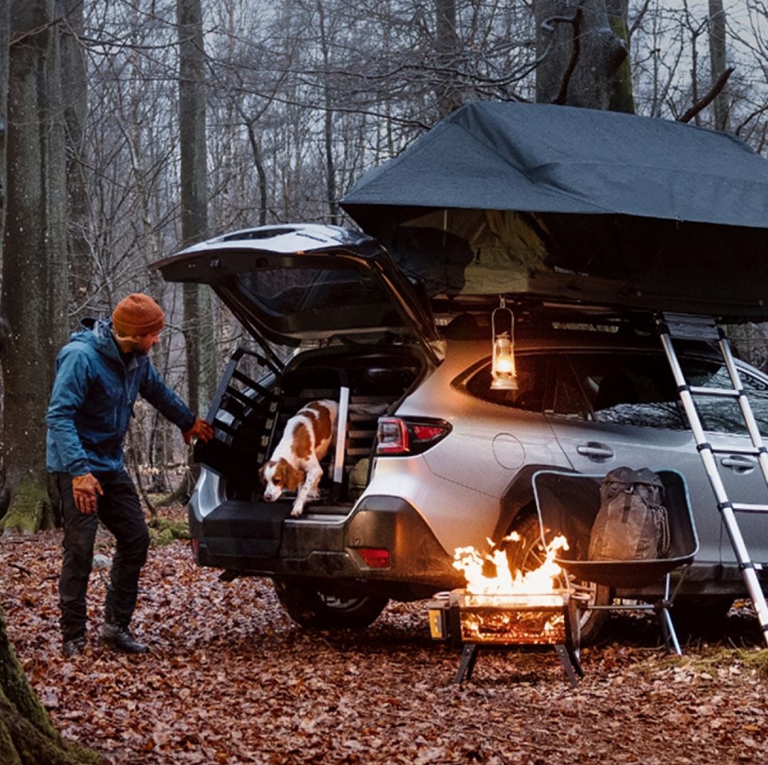 Persona acampando con su perro junto a un coche en el bosque, usando una jaula de viaje Thule Allax para perro para transporte seguro y protegido.