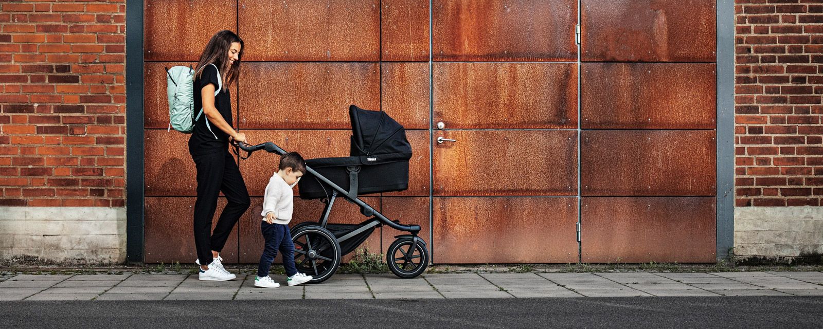 A woman walks with a Thule Urban Glide 2 bassinet stroller and her toddler walking beside her.