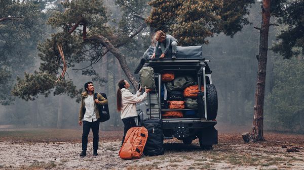 In a foggy forest, a family loads their duffel bags into a jeep with a rooftop tent on the roof.