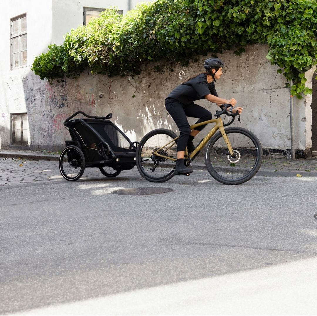 En kvinne sykler på en sykkel som trekker en Thule Chariot-sykkelhenger med en baby inni. 