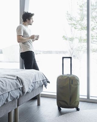 한 남성이 호텔 객실에서 커피를 들고 옆에 여행 가방을 둔 채 창밖을 내다보고 있습니다.