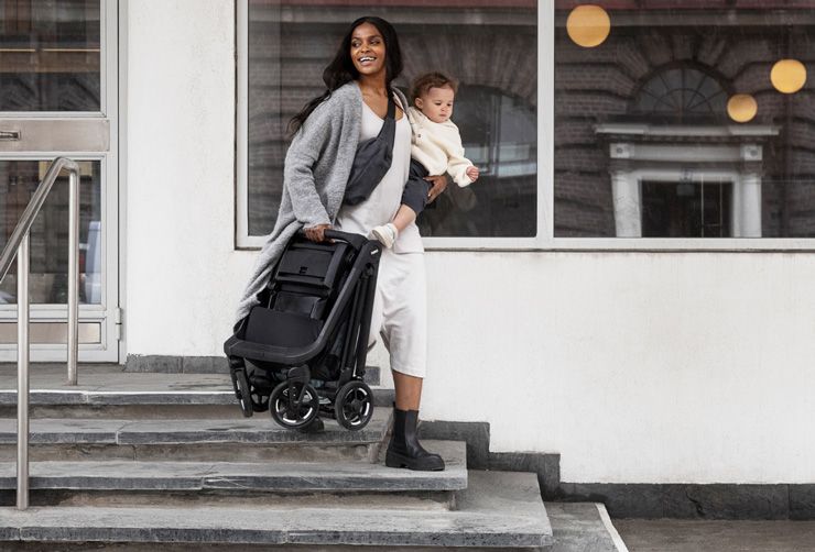 A mother walks down stairs holding a folded Thule Shine stroller in with one hand and a baby in the other.