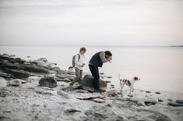  A man and a  woman are looking at their dog at the water’s edge.