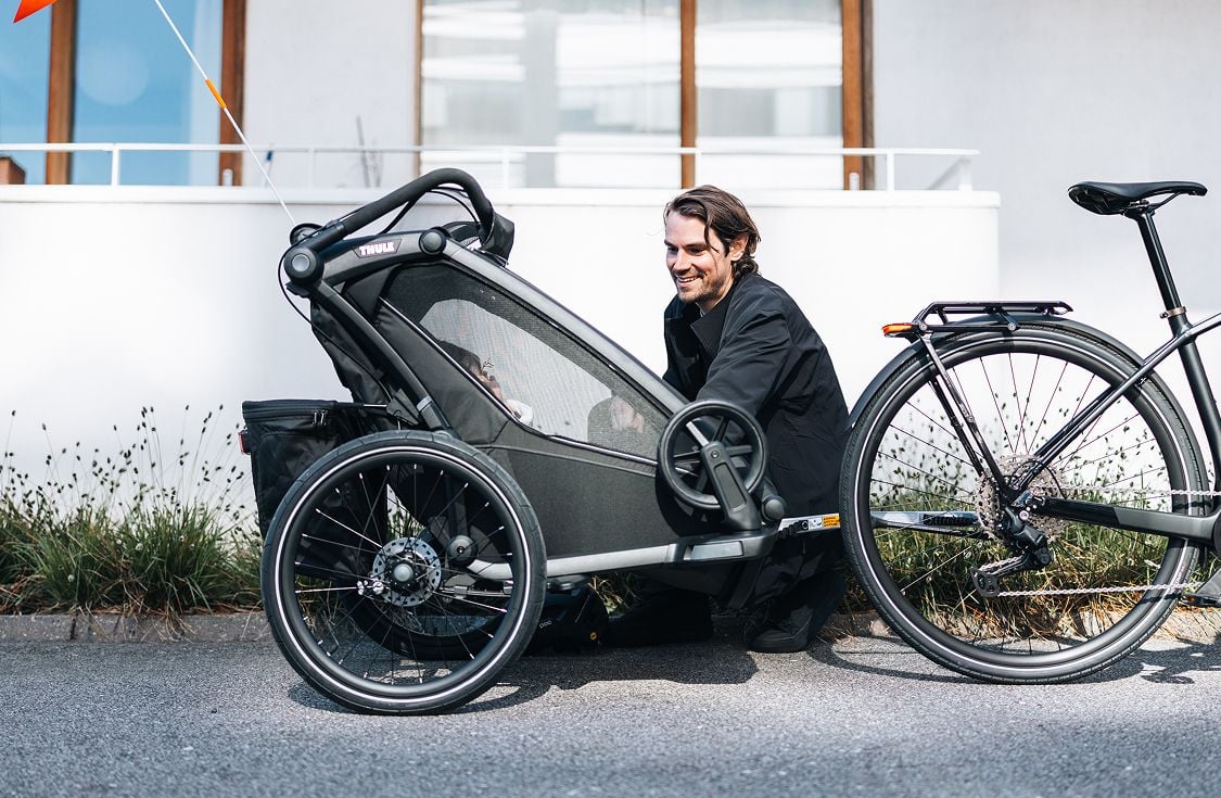 Uma pessoa se agacha ao lado de uma bicicleta com um trailer de bicicleta infantil acoplado ao longo de um caminho pavimentado ao lado de um prédio e de áreas baixas de verde.