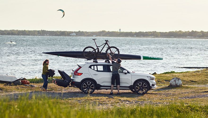 一辆白色 SUV 在海边行驶，车顶载有自行车和皮艇，车后安装有拖车箱。