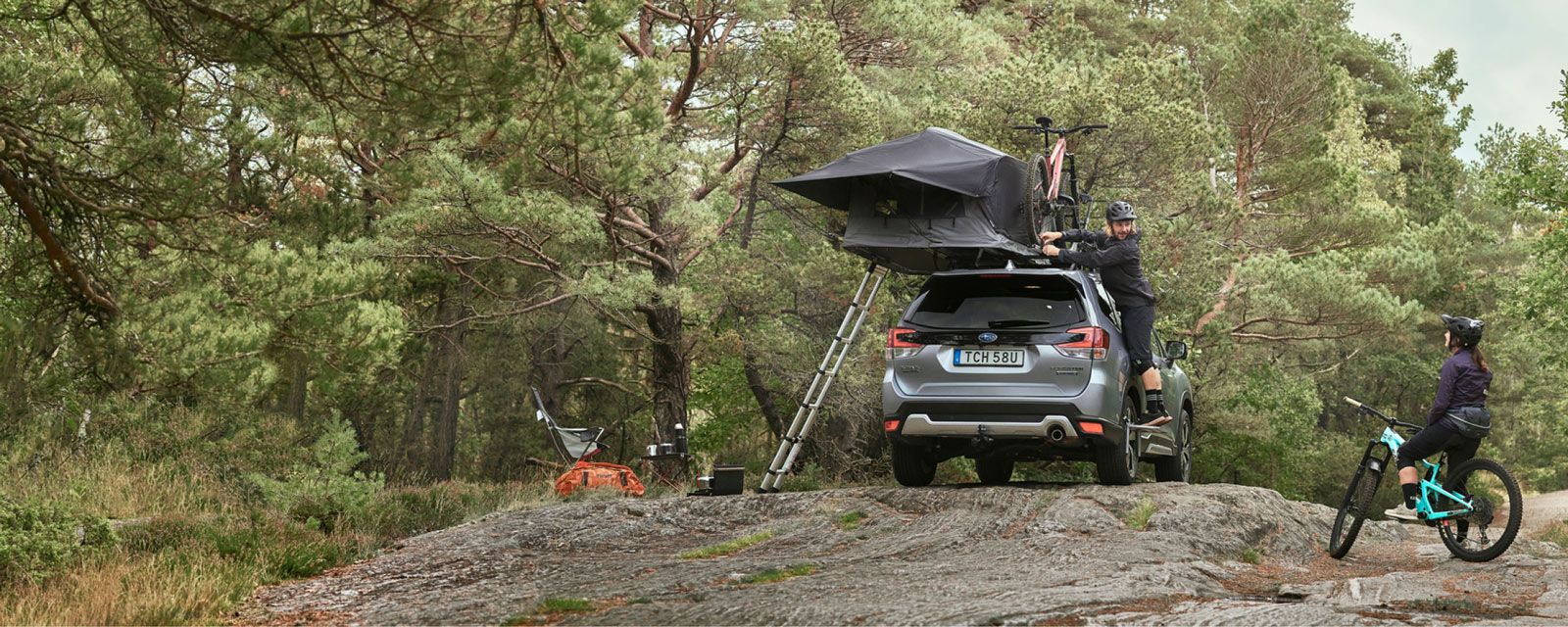 A man in a forest attaches the rain fly to the car roof tent on his car.