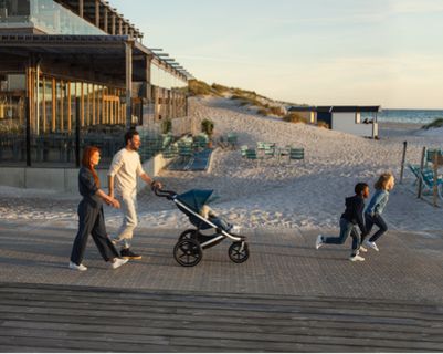 A man and woman holding hands are walking on a boardwalk while the man pushes a stroller and 2 kids run in front of them.