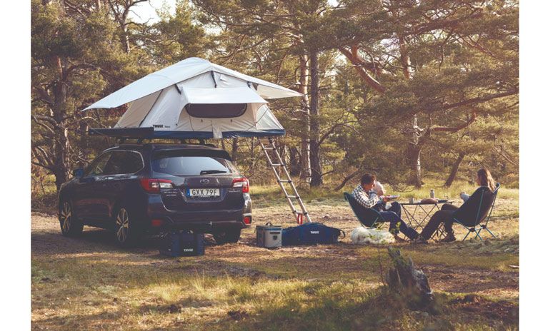 In a forest, two people sit on camp chairs beside their Thule Tepui Ayer rooftop tent.