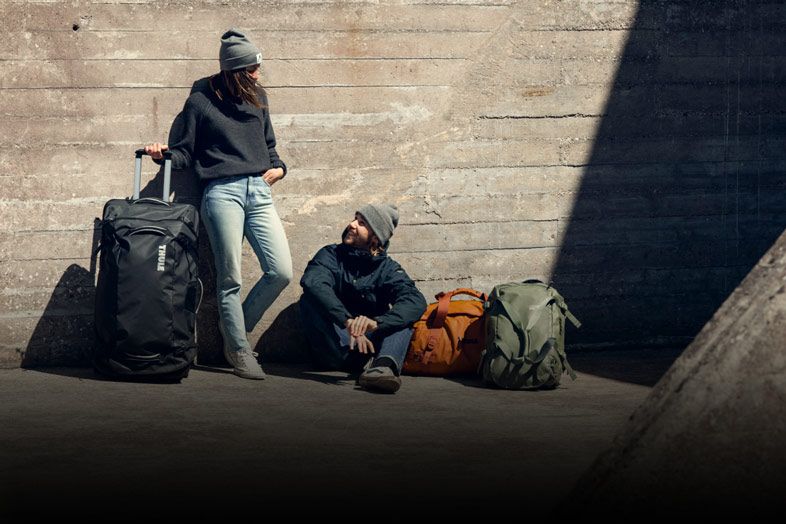 Two people lean against a wall with their Thule luggage.