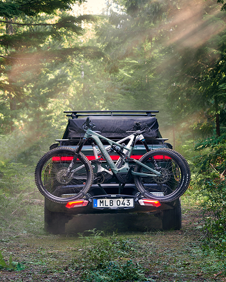 Un veicolo carico di due biciclette su un portabici Thule attaccato al retro si trova su un sentiero nel bosco. 