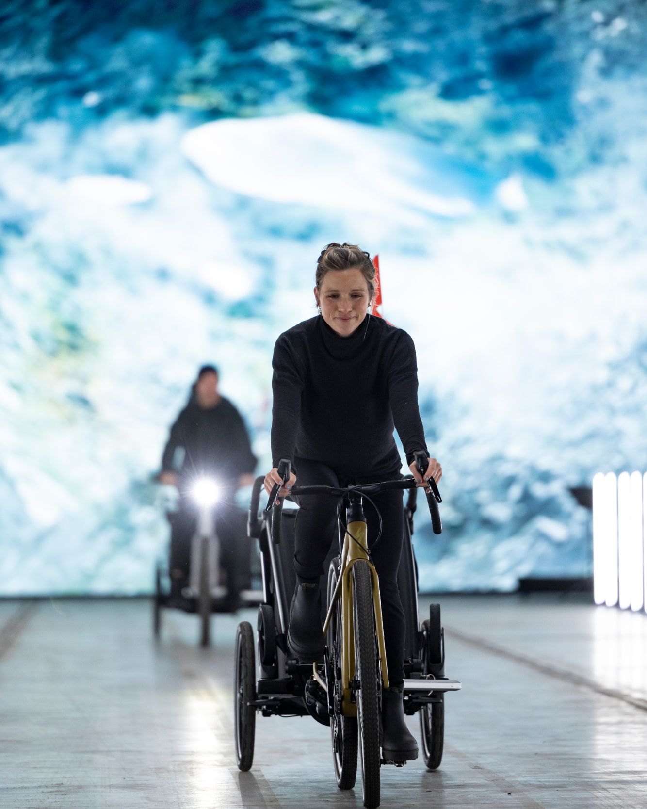 A woman wearing all black clothes riding a bicycle down a fashion runway with a Thule Chariot 3 bike trailer attached to the back. Behind her is another person blurred and also riding a bicycle down the same fashion runway.