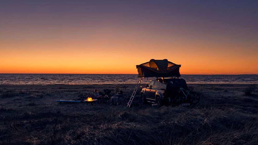 A vehicle is parked on the beach at sunset with a soft shell rooftop tent and people sitting around a camp fire.