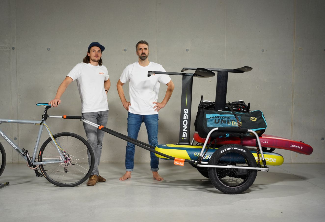 To menn poserer i et studio med en sykkel med Reacha Sport Beach-sykkelvogn med surfebrett og utendørsutstyr.