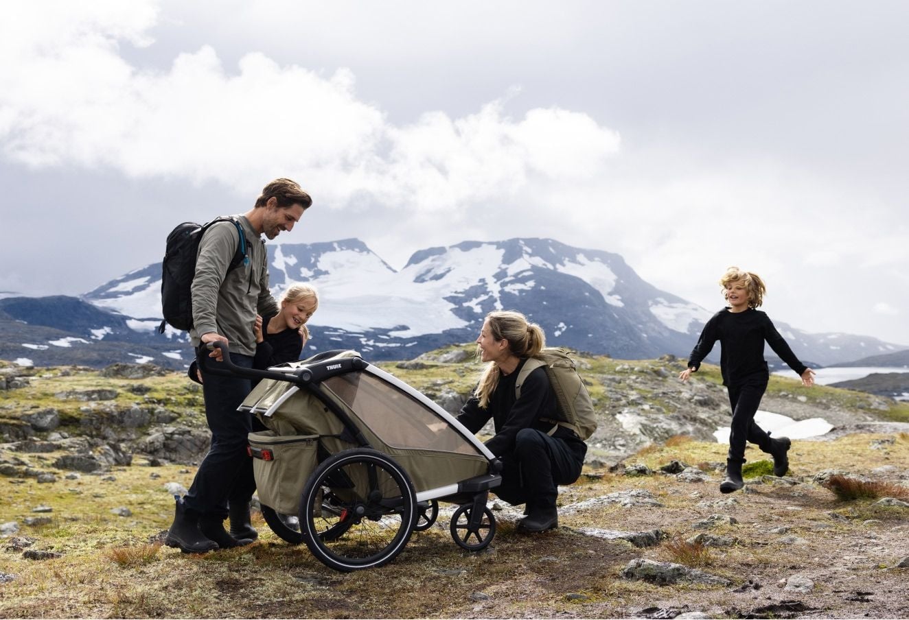 Família com dois filhos a usar um atrelado de bicicleta Thule Chariot enquanto caminham numa paisagem montanhosa.