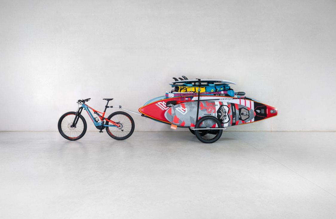 Una bicicleta estacionada en un estudio con un remolque de bicicleta Reacha Sport conectado en el que se transportan varias tablas de surf y SUP.
