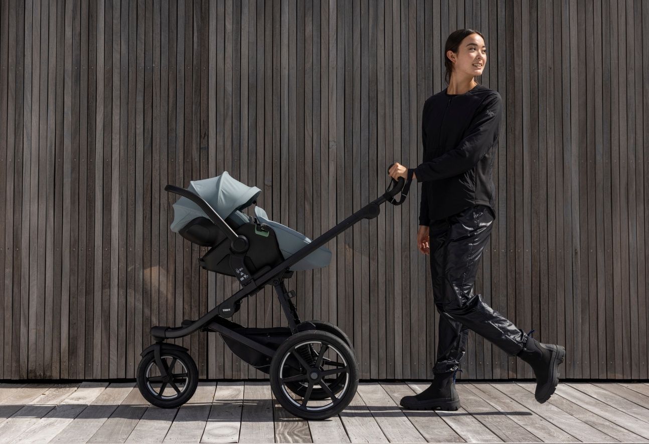 A woman with brown hair and black clothes walking with a Thule Urban Glide 3 stroller and Thule Maple infant car seat installed on it.