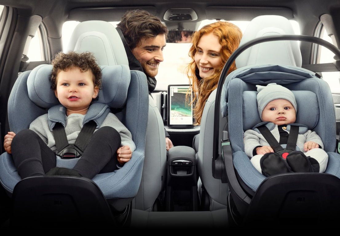 Two children sit in rear facing car seats while two smiling adults look back at them from the front seats of a vehicle.
