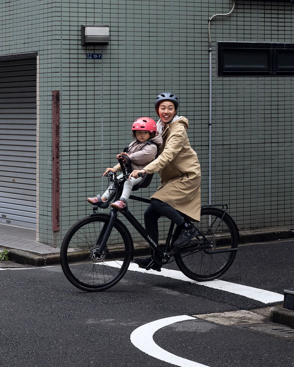 Adult riding a bicycle with a front-mounted child seat carrying a helmeted child, on a city street beside a tiled building.z