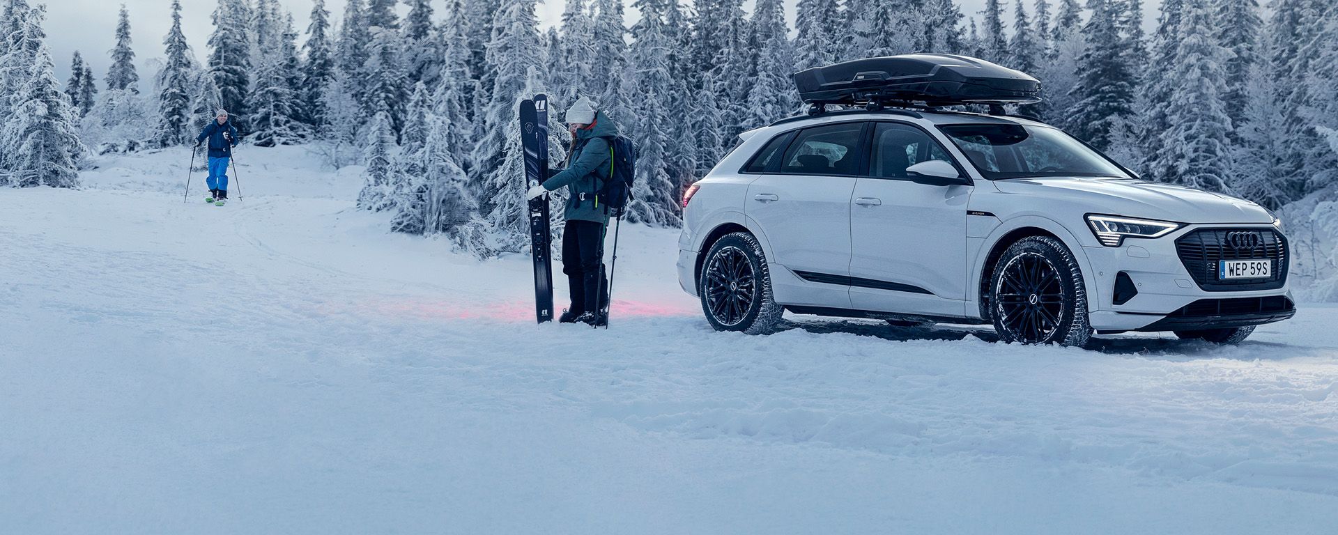 A car is parked by a snowy forest with a Thule Vector roof box, and skiers stand beside it.