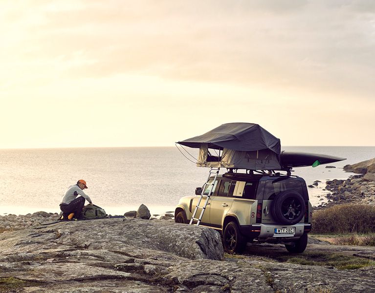 A man packs his duffel overlooking the water next to a car with a Thule Foothill rooftop tent