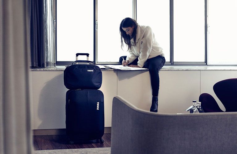 A woman sits on a windowsill, writing on a piece of paper next to two items of Thule luggage.