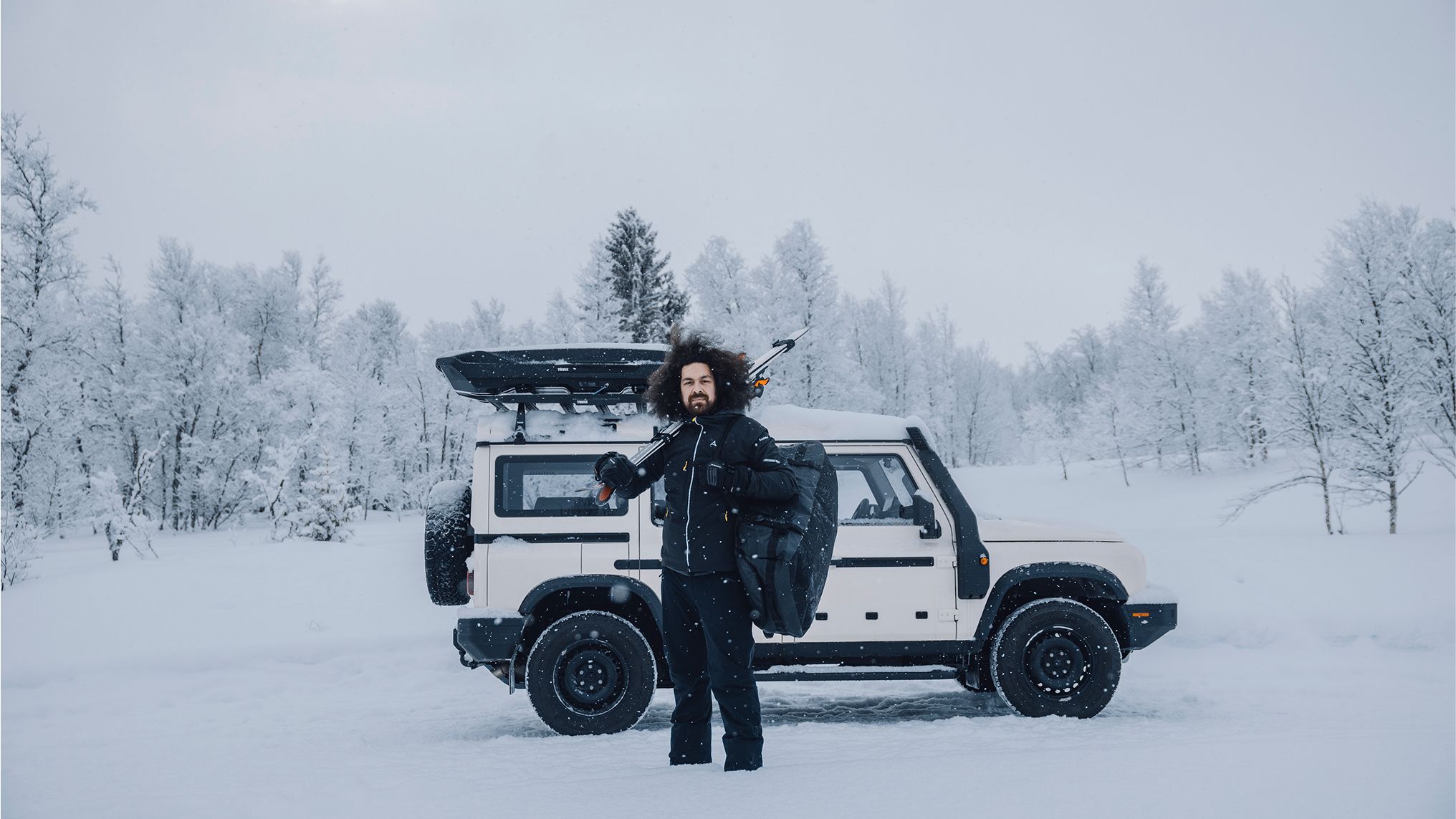 En person står foran en bil, der holder parkeret i snedækkede omgivelser. På taget af bilen er der en Thule Motion 3-tagboks. Personen bærer ski over højre skulder og har en sort Thule Chasm-rejsetaske over venstre skulder.