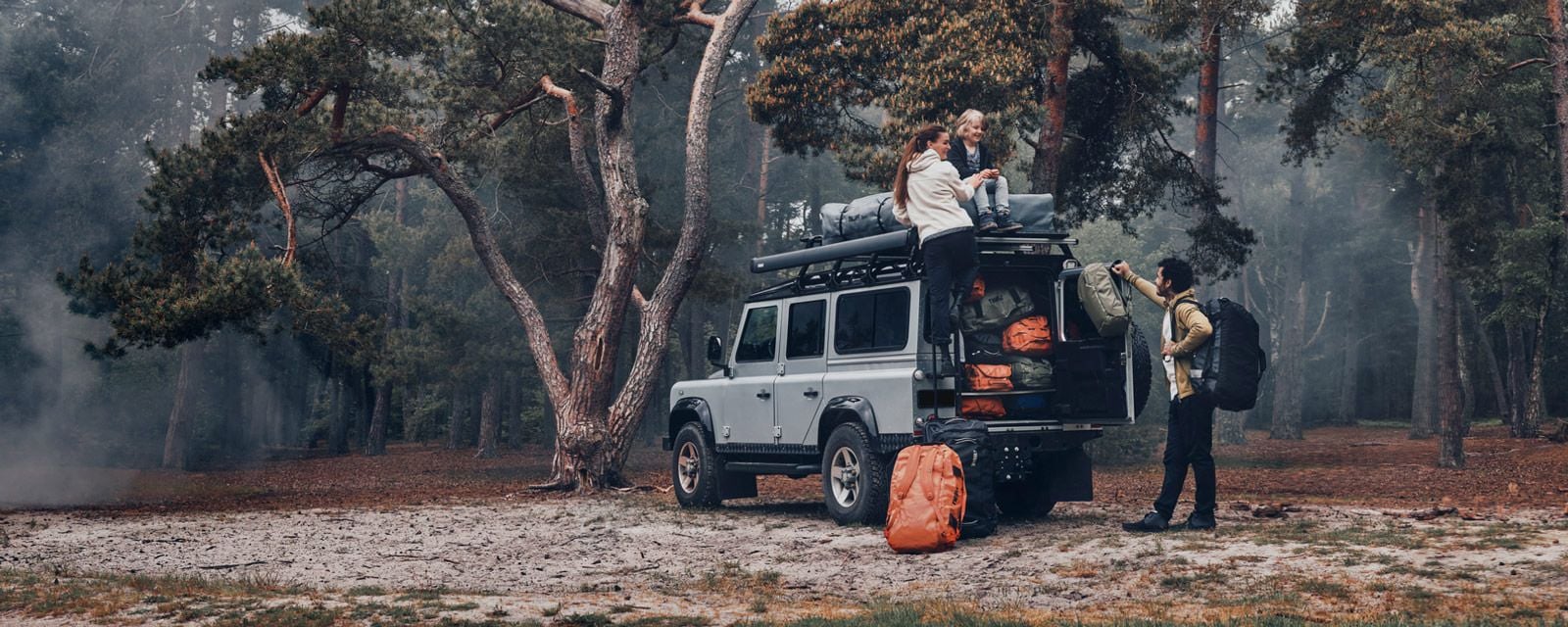 In a misty forest a family unloads Thule Chasm bags from their jeep which has a Thule rooftop tent on the roof.