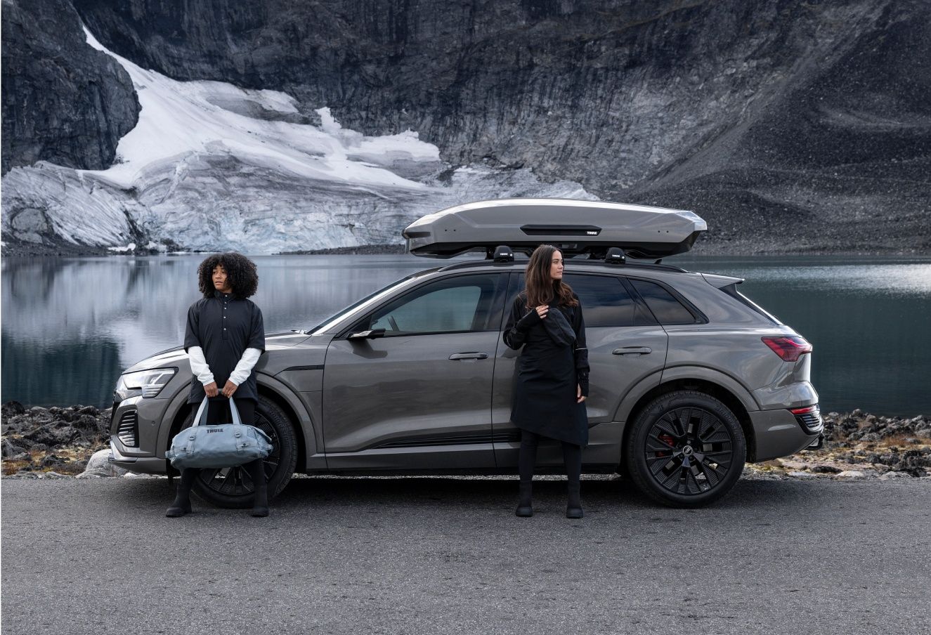 兩位女士，其中一人拿著 Thule Chasm 手提包，靠在一輛停在冰河前方、裝有 Thule Motion 3 車頂箱的灰色汽車上。