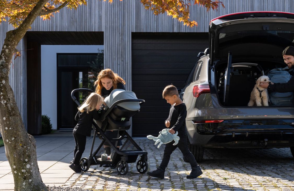 Mees seisab pagasiruumi juures, hoides koera nägu, samal ajal kui naine ja kaks last seisavad auto kõrval kärule paigaldatud Thule Maple laste turvaistme juures.