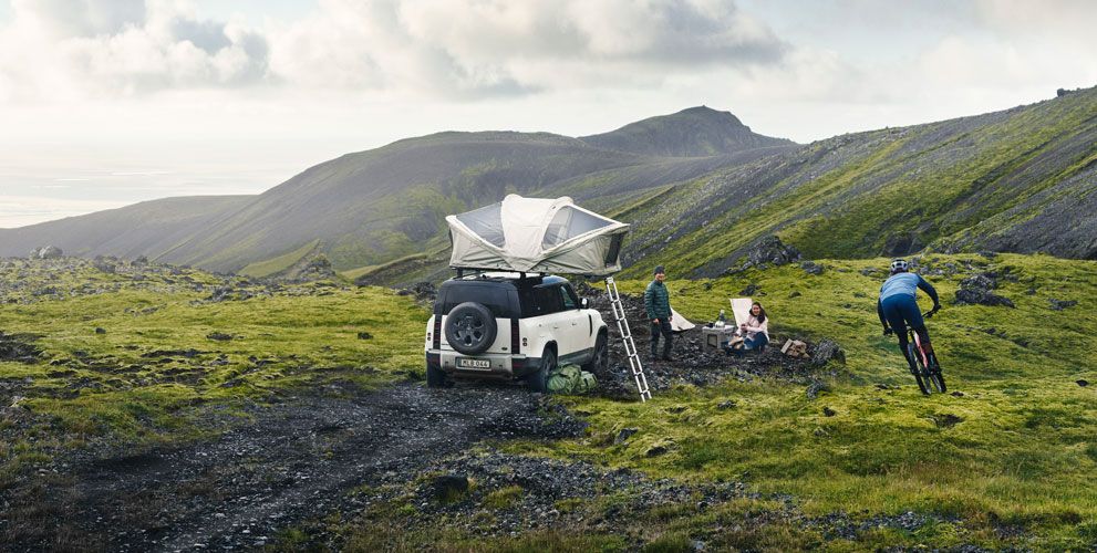 In a grassy misty field a vehicle is parked with a white Thule Approach rooftop tent and people sit with camp gear.