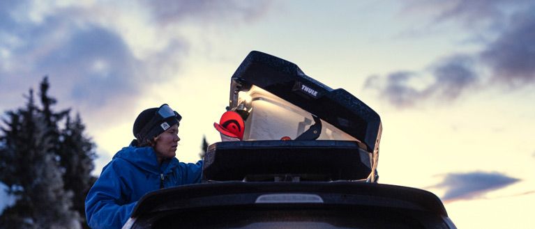 A close-up of a skier opening their Thule rooftop cargo carrier in a forest at sunset.