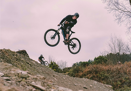 Ein Mann macht einen Fahrradtrick, während er durch die Luft fliegt, mit felsigem Gelände darunter. 
