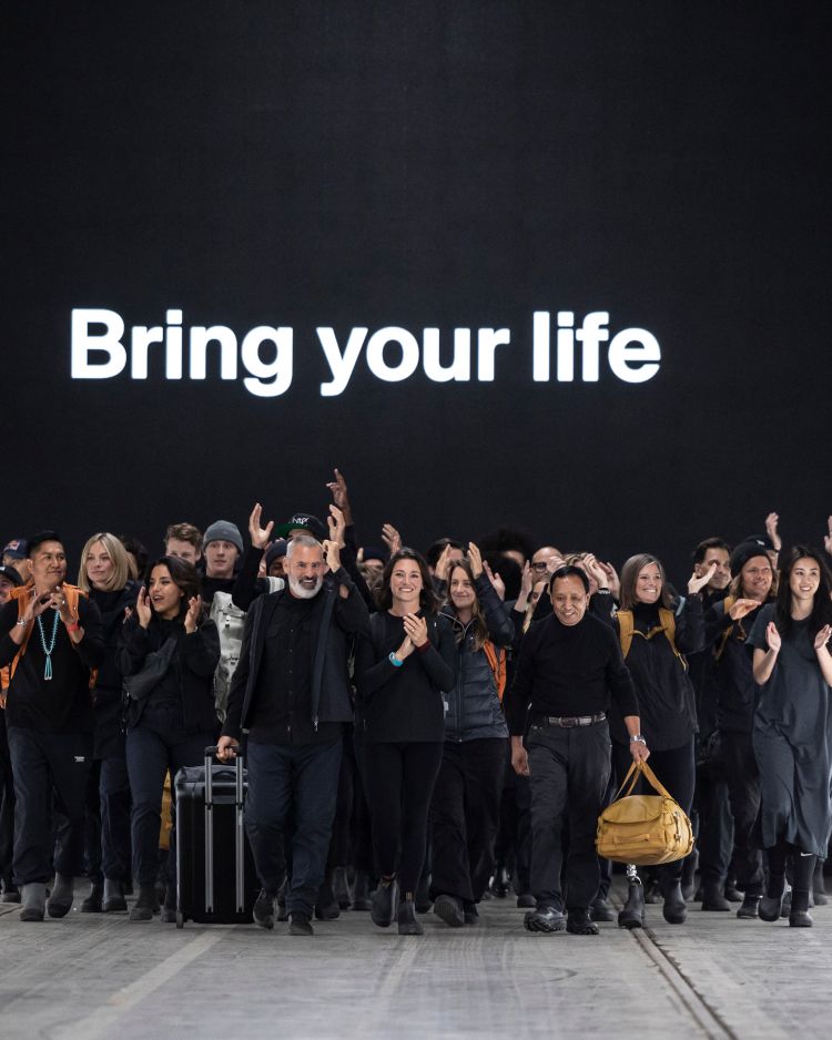 A big group of people all dressed in black looking happy walking down a fashion runway holding various Thule bags. Behind them is a text on the screen that says “Bring your life”.