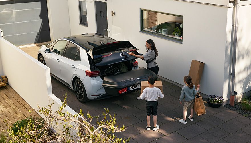 Uma mulher carrega itens no porta-malas de um carro enquanto seus filhos ajudam a carregar itens na caixa traseira da barra de reboque.