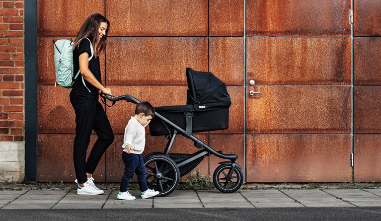 A woman walks with a Thule Urban Glide 2 bassinet stroller and her toddler walking beside her.