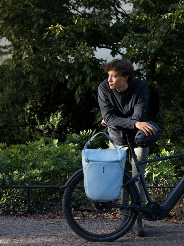 En mand, en sort cykel og en blå taske foran grønne træer.