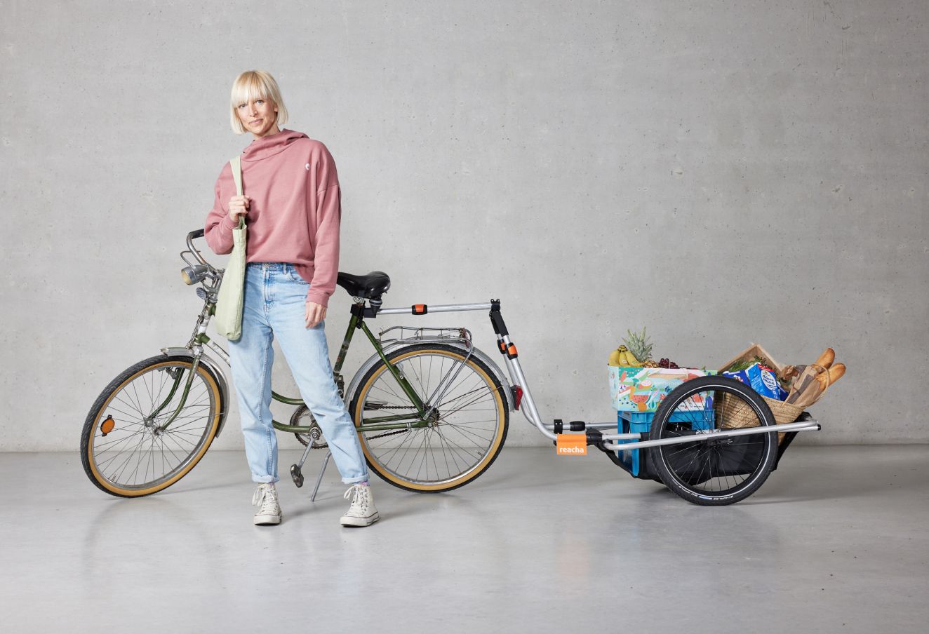 Kobieta stoi w studio z przyczepką rowerową Reacha City przymocowaną do roweru, na przyczepce artykuły spożywcze i inne przedmioty.