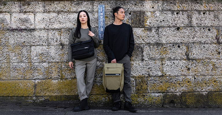 Two people stand with their back to a mossy wall holding the Thule Paramount crossbody and backpack.