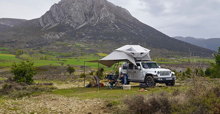 两人坐在配有 Thule Approach 车顶帐篷的吉普车遮阳篷下的野营折叠椅上，吉普车停在山间。