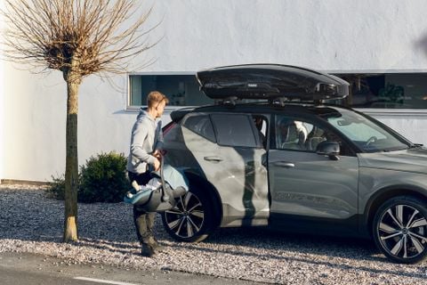 Un uomo che sta in piedi accanto a un'auto parcheggiata vicino ad una casa bianca, si appresta a caricare un seggiolino per auto Thule Maple sul sedile posteriore.