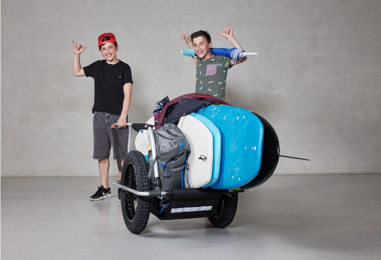 Dos jóvenes sonrientes en un estudio que muestra un remolque de bicicleta Reacha Sport Beach cargado con una gran tabla de paddleboard azul y equipo de acampada.