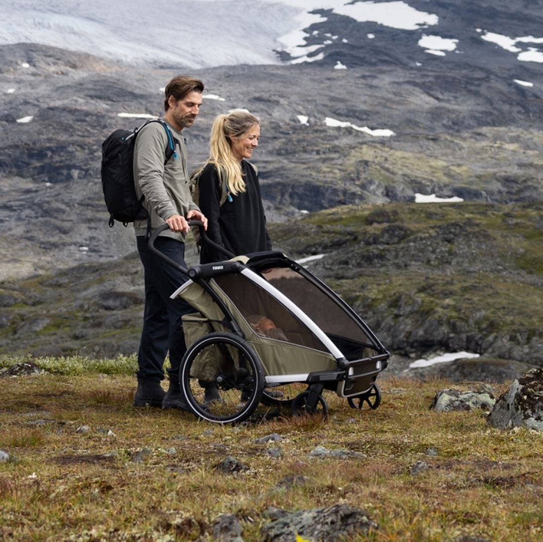 Med en bergsbakgrund står två vuxna nära en Thule-barnvagn med en bebis i medan två barn leker i närheten.
