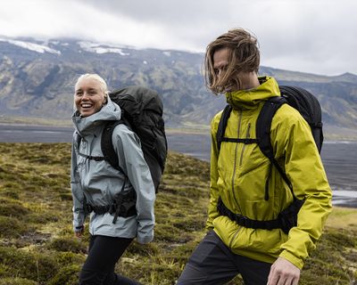 Dos personas paseando por un campo junto a una montaña con mochilas de senderismo.